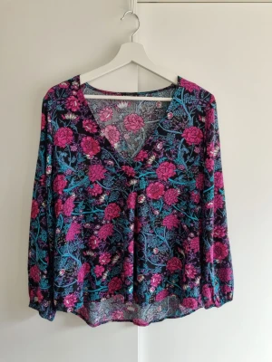 Blommig långärmad blus Medicine S - Blus med färgstarkt blommönster i lila, rosa och turkos. V-ringad framtill och avslappnad passform. Långa ärmar med resår vid ärmslut. 100% viskos, perfekt skick.
