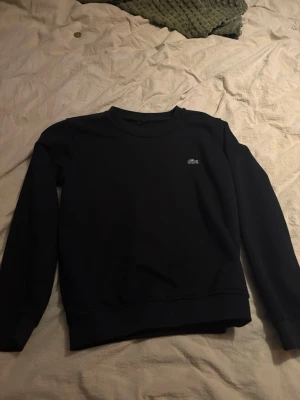 Svart sweatshirt från Lacoste - Klassisk svart sweatshirt från Lacoste med rund halsringning och diskret krokodillogga på bröstet. Tröjan har långa ärmar och ribbade muddar vid ärmslut och nederkant. Perfekt för en clean och stilren look.