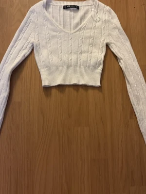 Vit croppad kabelstickad tröja - Säljer en vit croppad kabelstickad tröja från pull & bear. Tröjan har v-ringning, långa ärmar och bred ribbad mudd i midjan. Perfekt för dig som gillar trendiga och bekväma plagg med snygg passform.