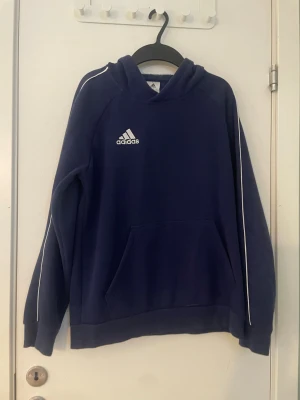 Mörkblå hoodie från Adidas - Snygg mörkblå hoodie från Adidas med vit logga på bröstet och vita detaljer längs ärmarna. Klassisk känguruficka framtill och huva. Perfekt för en sportig och avslappnad stil.