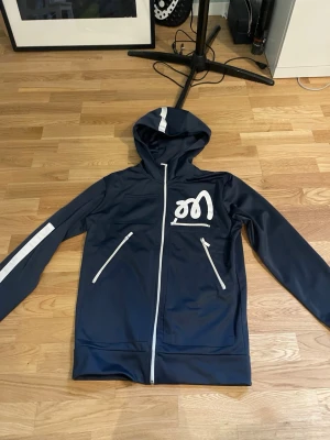 Marinblå hoodie med dragkedja och tryck - Snygg marinblå hoodie med vit dragkedja, vita detaljer och ett tryck på bröstet. Huva med vit rand och två dragkedjefickor framtill. Tillverkad i ett mjukt och sportigt material, perfekt för en avslappnad stil.