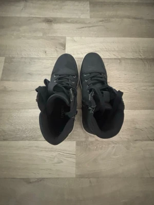 Svarta high top sneakers med dragkedja - Snygga svarta high top sneakers med vita sulor och bruna yttersulor. Skorna har både snörning och dragkedja på sidan för enkel påtagning. Materialet ser ut att vara en mix av textil och syntet. Perfekta för dig som gillar streetwear och vill ha något stilrent.