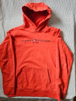 Röd hoodie från Tommy Hilfiger - Snygg röd hoodie från Tommy Hilfiger med broderad logga och text på bröstet. Klassisk känguruficka framtill och ribbade muddar vid ärmar och nederkant. Tillverkad i mjuk bomullsmix och har en huva för extra komfort. Perfekt för en avslappnad stil.