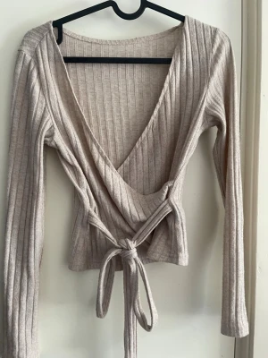 Beige ribbad omlottopp med knyt - Trendig beige omlottopp med ribbad struktur och långa ärmar. Toppen har djup v-ringning och knytband i midjan för en snygg passform. Perfekt att styla med jeans eller kjol för en cool look.
