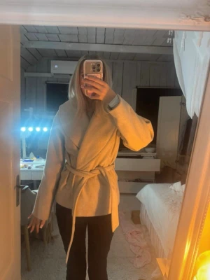 Beige kort kappa - Stilren beige kappa med knytskärp i midjan och bred krage. Kappan har en rak passform och långa ärmar, perfekt för lager på lager under kyliga dagar. Materialet ser ut att vara mjukt och bekvämt, vilket ger en avslappnad men ändå chic look. Storlek m men passar mig som har s. Den är lite nopprig