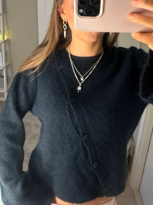 Mörkblå kofta med knappar - Snygg mörkblå kofta med asymmetrisk knäppning framtill och stora muddar vid ärmsluten. Koftan har en avslappnad passform och är gjord i ett mjukt, stickat material som passar perfekt till jeans eller kjol.