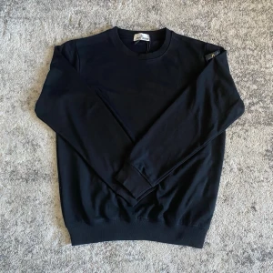 Svart stickad tröja från Stone Island - Snygg svart stickad tröja från Stone Island med rund halsringning och ribbade muddar. Tröjan har den klassiska Stone Island-loggan på vänster ärm och är perfekt för en clean och stilren look.