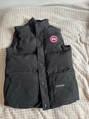 Svart dunväst från Canada Goose - Snygg svart dunväst från Canada Goose med hög krage och två stora fickor framtill. Västen har en klassisk Canada Goose-logga på bröstet och stängs med tryckknappar. Perfekt för lager-på-lager och håller dig varm under kyliga dagar. 