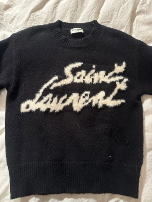 Svart Saint Laurent stickad tröja - Stickad svart tröja från Saint Laurent med vit fuzzy logga framtill i graffiti-stil. Tröjan har rund halsringning, ribbade muddar och nederkant. Materialet ser mjukt och lite luddigt ut, perfekt för en trendig och avslappnad look. Använd i en månad. 