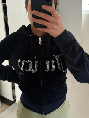 Äkta Juicy Couture svart velour hoodie - Juicy Couture hoodie i svart velour med dragkedja framtill och huva. Stora glittriga stenar bildar Juicy-loggan på bröstet. Mjuk och skön känsla, ribbade muddar och fickor framtill. Perfekt för en chill och trendig look.