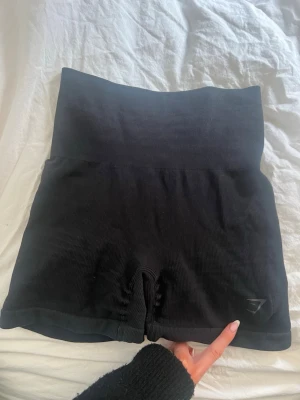 Svarta ribbade shorts från Gymshark - Säljer ett par svarta ribbade shorts från Gymshark med hög midja och tight passform. Perfekta för träning eller chill dagar. Stretchigt material som sitter skönt och formar sig efter kroppen. Diskret Gymshark-logga på benet.