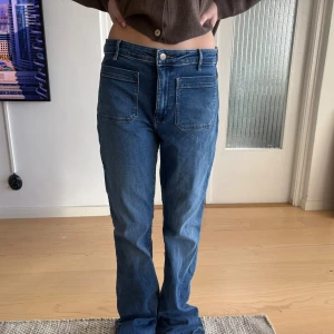 Wrangler jeans med fickor fram - Säljer ett par blå jeans med flare ben och stora fyrkantiga fickor framtill. De är utsvängda men väldigt stora på mig så det syns ej. Jeansen har hög midja och klassisk knappgylf. Materialet är denim och färgen är en klassisk mellanblå tvätt. Perfekta för en avslappnad och trendig look. Knappt använda. Köpte för 999kr. 