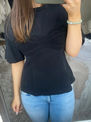 Svart topp med virkad detalj - Snygg svart topp med unik virkad detalj över bysten. Modellen har korta ärmar och en figurnära passform som framhäver siluetten. Perfekt att styla med jeans eller kjol för en trendig look.