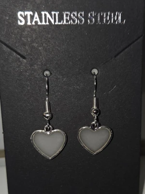 Hjärthängande örhängen i stål  - Snygga örhängen i rostfritt stål med hjärtformade hängen. Hjärtana har en skimrande yta som skiftar mellan vitt och lila beroende på ljuset. Klassisk krok för enkel användning och stilren metallram runt hjärtat. Tål vatten & missfärgas inte. Lång livslängd.