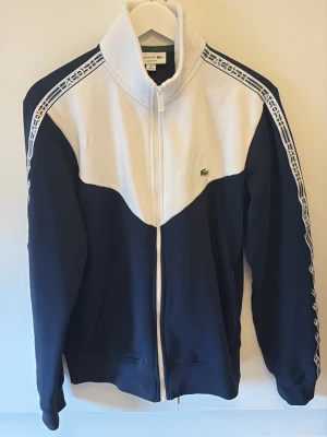 Blåvit ziptröja från Lacoste - Sportig blå och vit ziptröja från Lacoste med hög krage och logga på bröstet. Tröjan har vita detaljer längs ärmarna med Lacoste-tryck och är tillverkad i mjukt material. Perfekt för en avslappnad och trendig look.
