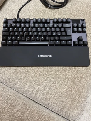 SteelSeries Apex Pro TKL Gen 3 - Säljer ett svart mekaniskt tangentbord från SteelSeries med avtagbart handledsstöd och USB-kabel. Tangentbordet har kompakt design och tydliga vita tecken på knapparna, perfekt för gaming eller arbete vid datorn.
