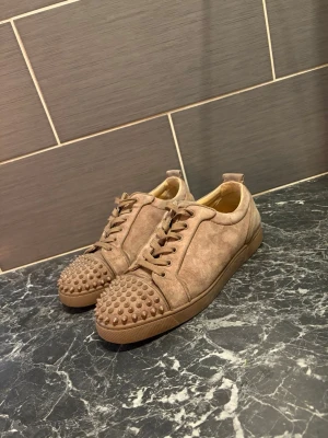 Christian Louboutin skor - Snygga beige sneakers från Christian Louboutin i mocka med ikoniska röda sulor och coola nitar på tån. Lågt skaft och klassisk snörning. Pris kan diskuteras 