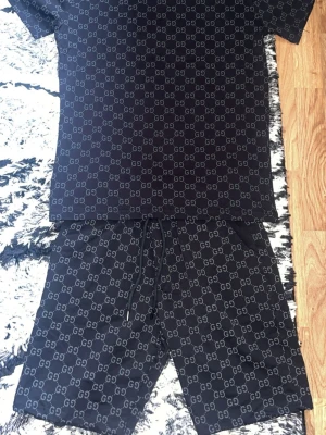 Svart GG-mönstrat set från Gucci - Snyggt svart set från Gucci med all-over GG-logga. Setet består av en t-shirt och matchande shorts med dragsko i midjan. Materialet ser mjukt och bekvämt ut, perfekt för en chill look. Passformen är avslappnad och mönstret ger en exklusiv vibe.