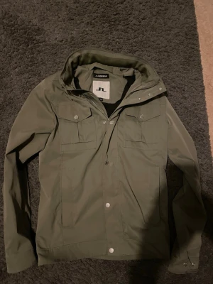 Olivgrön vindjacka från J.Lindeberg - Snygg olivgrön vindjacka från J.Lindeberg med huva, dragkedja och tryckknappar. Jackan har två bröstfickor med lock och två sidofickor. Perfekt för blåsiga dagar och enkel att matcha med olika outfits.