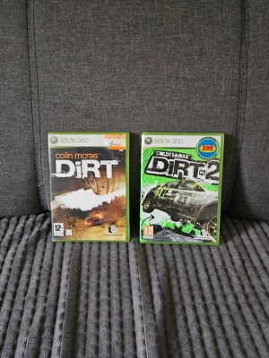 Colin McRae DiRT 1 & 2 Xbox 360 spel - Två rallyspel till Xbox 360: Colin McRae DiRT och DiRT 2. Båda spelen har gröna plastfodral med färgstarka omslag och racingbilar i fokus. Perfekt för dig som gillar fartfyllda offroad-race och klassiska rallybanor.