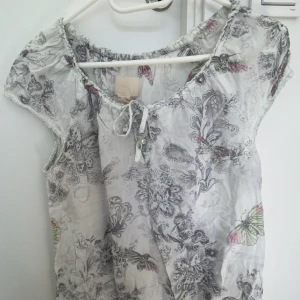 Vit blus med mönster och knytning - Säljer en söt vit blus med vitt/grått och fjärilsmönster. Blusen har korta ärmar, rundad halsringning med knytning framtill och är tillverkad i ett tunt, luftigt material. Finns inte längre och köpa, Perfekt för sommaren!
