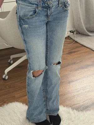 Ljusblå slitna bootcut jeans - Säljer ett par ljusblå bootcut jeans med slitningar och hål på knäna. Jeansen har klassisk femficksdesign, knappstängning och bakfickor med lock och knapp. Perfekta för en avslappnad och trendig look.