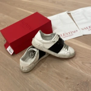 Valentino sneakers med svart detalj - Säljer ett par vita Valentino sneakers med bred svart rem över mitten. Skorna har rund tå och platt sula, samt diskreta nitar på hälen. Klassisk och stilren design med tydlig logga på sidan. Köp idag 1100 annars fast på 1500