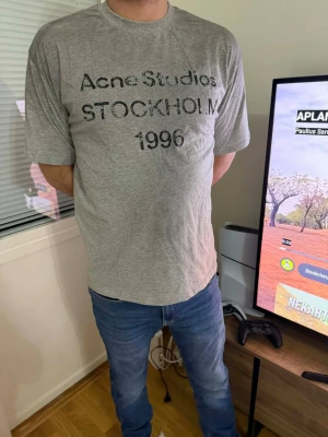 Grå Acne Studios t-shirt - Snygg grå t-shirt från Acne Studios med trycket 'Acne Studios STOCKHOLM 1996' på bröstet. Klassisk passform med rund hals och korta ärmar. Perfekt basplagg för en clean och avslappnad stil.