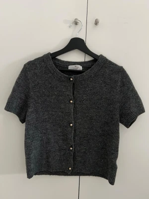 Mörkgrå stickad kortärmad kofta från H&M med guldknappar - Kortärmad kofta i mörkgrått från H&M med rund halsringning och ribbade kanter. Koftan har dekorativa guld knappar framtill och är stickad i ett mjukt material som ger en avslappnad look. Perfekt att slänga över en topp för en enkel och snygg stil. Använd 2-3 gånger men matcha inte min stil, tyvärr. Annars jätte snygg och top kvalitet❤️