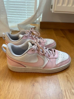 Nike rosa och vita låga sneakers - Snygga sneakers från Nike i en fräsch kombination av ljusrosa och vitt. Skorna har klassisk låg siluett, perforerad tåbox för ventilation och matchande rosa skosnören. Tillverkade i syntetmaterial och textilfoder för skön komfort. Perfekta för en trendig vardagslook.