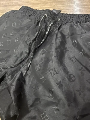 Svarta LV bad shorts - Snygga svarta shorts från LV. Shortsen har elastisk midja med snörning där loggan syns på banden. Tillverkade i ett glansigt syntetmaterial som ger en exklusiv känsla.