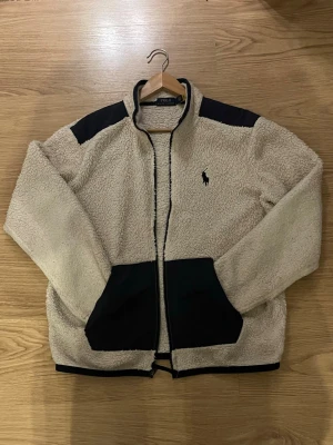 Beige teddyjacka från Polo Ralph Lauren - Mysig teddyjacka i beige från Polo Ralph Lauren med svarta detaljer på axlar och fickor. Dragkedja framtill och broderad logga på bröstet. Perfekt för kalla dagar och riktigt snygg till jeans eller mjukisbyxor.