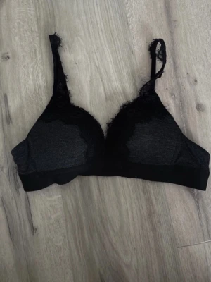 Svart spets-bh med justerbara band - En svart bh med mjuka kupor och spetsdetaljer längs kanterna. Bh:n har justerbara axelband och hakknäppning bak. 