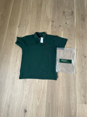 Mörkgrön pikétröja från Polo Ralph Lauren - Klassisk mörkgrön pikétröja från Polo Ralph Lauren med korta ärmar och krage. Tröjan har den ikoniska röda logobroderingen på bröstet och är tillverkad i mjuk bomull. Perfekt till jeans eller shorts för en avslappnad look.