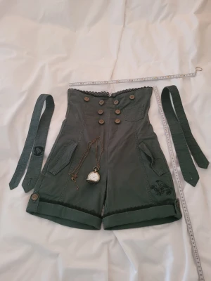 Steampunk shorts Mad Hatter cosplay - Unika gröna steampunk-inspirerade shorts med hög midja, dekorativa knappar och spetsdetaljer. Medföljer två matchande band och en Mad Hatter-fickur i metall. Perfekt för cosplay eller maskerad med Alice i Underlandet-tema.