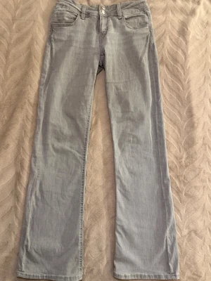Ljusgrå lågmidjade bootcut jeans med dubbla knappar - Säljer ett par ljusgrå jeans med bootcut passform och dubbla knappar i midjan. Jeansen har klassiska fem fickor och bälteshällor. Materialet är mjukt denim som ger en avslappnad look. Perfekta för dig som gillar en enkel men snygg stil. Stretchig matril väldigt bekväma.