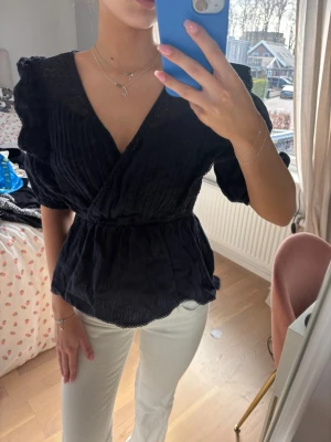 Mörkblå omlottblus med spetsdetaljer - Trendig svart omlottblus med v-ringning och fina spetsdetaljer längs halsringningen. Blusen har puffiga korta ärmar och markerad midja som ger en snygg siluett. Materialet ser lätt och luftigt ut, perfekt för en stilren look.