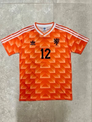 Holland retro fotbollströja Adidas - Orange retro fotbollströja från Adidas med geometriskt mönster, svarta detaljer och nummer 12. På bröstet finns Adidas-logga och Hollands landslagsemblem, på ryggen står det 'Van Basten' och nummer 12. Klassisk v-ringad krage och vita ränder på axlarna.