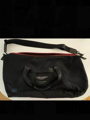 Aston Martin Racing (Hackett) svart weekendbag - Snygg svart weekendbag, HACKETT x Aston Martin Racing med justerbar axelrem och dubbla handtag. Väskan har röd dragkedja, diskret logga framtill och en praktisk ytterficka med dragkedja på baksidan. Tillverkad i slitstarkt tygmaterial och passar perfekt för resor eller gymmet. Mått CA: Längd: 55cm. Bredd:25cm. Höjd: 25cm