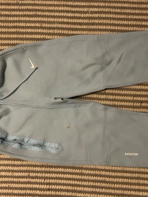 Ljusblå mjukisbyxor från Nike NOCTA - Snygga ljusblå mjukisbyxor från Nike NOCTA med dragsko i midjan och ribbade benslut. Byxorna har diskreta fickor på sidorna och vita loggor på benet och vid höften. Perfekta för en chill och avslappnad stil.