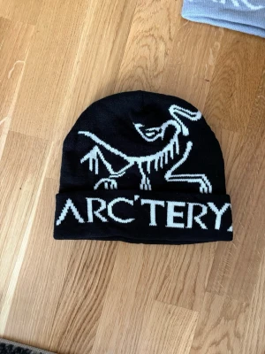 Svart mössa från Arc'teryx - Säljer en svart mössa från Arc'teryx med vit logga och text framtill. Mössan är ribbstickad med uppvikt kant och har en stilren design. Perfekt för dig som vill ha en snygg och funktionell accessoar till vintern.