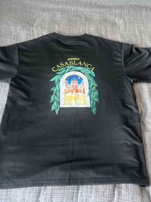 Svart t-shirt med fina detaljer - Oanvänd‼️Perfekt för sommaren, Snygg svart t-shirt från Ave/nida med färgglatt tryck av Casablanca och ett motiv av en byggnad omgiven av gröna blad. T-shirten har rund hals och korta ärmar, perfekt för dig som gillar grafiska prints och streetstyle.