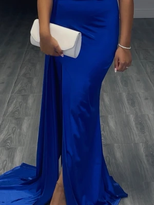 Kornblå långklänning med slits - Säljer en elegant kornblå långklänning i glansigt material med hög slits framtill. Klänningen har en figurnära passform och ett långt släp som ger en dramatisk look. Perfekt för speciella tillfällen när du vill sticka ut.endast använd 1 gång.