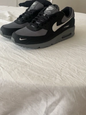 Nike Air Max 90 svart/grå sneakers - Nike Air Max 90 sneakers i svart och grått med vit swoosh. Klassisk siluett med synlig Air Max-dämpning i sulan. Ovandel i mesh och mocka för både stil och komfort. Grå yttersula med tydligt mönster för bra grepp. Perfekta för en sportig och trendig look.