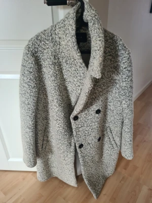 Gråmelerad kappa från Vila - Snygg gråmelerad kappa från Vila med dubbelknäppning och bred krage. Kappan har två fickor framtill och är tillverkad i ett mjukt, ull-liknande material. Perfekt för kyliga dagar och ger en trendig look till din outfit.