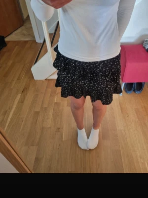 Svart blommig volangkjol - Säljer en svart kortkjol med små vita blommor och volangdetaljer. Den har resår i midjan och passar till det mesta, jättesnygg på sommaren