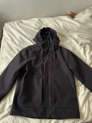 Svart regnjacka med huva - Snygg svart regnjacka med justerbar huva och dragsko. Jackan har två stora framfickor och stängs med dragkedja och tryckknappar. Materialet är vattenavvisande och jackan har en stilren, minimalistisk design som passar perfekt till regniga dagar.