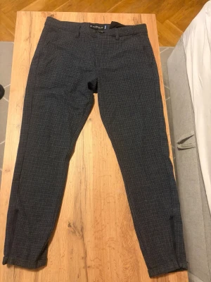 Mörkgrå rutiga kostymbyxor Gabba - Snygga mörkgrå kostymbyxor från Gabba med diskret rutmönster. Byxorna har smal passform, bälteshällor, sidofickor och två bakfickor. Perfekta för en stilren look och passar till skjorta eller t-shirt.