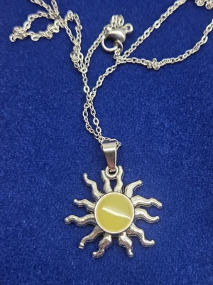 Solhalsband med färgskiftande hänge - Unikt halsband med ett solformat hänge i silverfärgad metall. Hängets mitt är en rund pärlliknande sten som skiftar färg mellan gult och blått beroende på ljus. Kedjan är tunn i rostfritt stål och har ett klassiskt lås. Perfekt för dig som gillar statement-smycken med magisk känsla. Vanligt dagsljus aktiverar effekten i hänget och det börjar lysa blått i mörker. Handgjord, tål vatten & har en lång livslängd. Kedjans längd: 45 cm, hänget: ca 2,5 - 3 cm. 