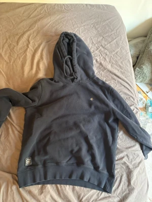 Mörkgrå hoodie från Morris Stockholm  - Snygg mörkgrå hoodie från Morris med klassisk huva och dragsko. Diskret Morris-logga på bröstet och en större patch vid nederkanten. Tillverkad i mjukt material som känns skönt mot huden. Perfekt för en avslappnad och stilren look.